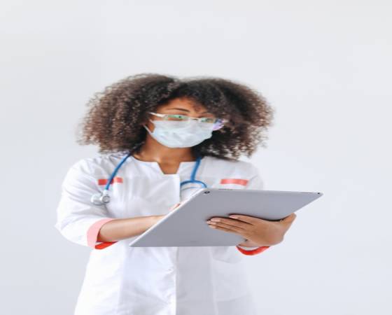 Home A female doctor wearing a mask uses a tablet in a medical setting, exemplifying modern healthcare.